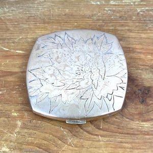 Puede incluir: Un estuche compacto de plata con un diseño floral grabado en la superficie.