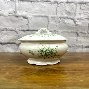 May include: A white ceramic lidded jar with a green floral design. The jar has a scalloped edge and a simple handle.