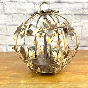 May include: A white metal chandelier with a round, cage-like design. The chandelier has a distressed finish and features leaf-shaped accents. It has four light sockets.