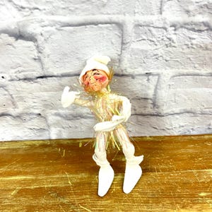 May include: A white elf figurine with a white hat and iridescent tinsel hair. The elf is smiling and has rosy cheeks. The elf is standing on a wooden surface.