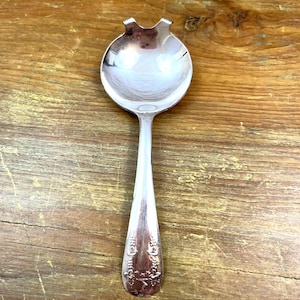 May include: A silver serving spoon with a rounded bowl and a decorative floral pattern on the handle. The spoon has a unique notched design on the bowl's edge. The spoon is made of a shiny, reflective metal.