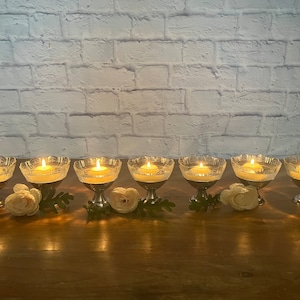 Puede incluir: Ocho candelabros de vidrio transparente con bases plateadas y velas encendidas. Los candelabros están dispuestos en fila sobre una superficie de madera.