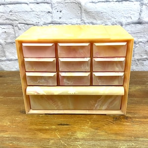 Puede incluir: Un joyero vintage con una combinación de colores melocotón claro y rosa. La caja tiene nueve cajones pequeños y un cajón inferior más grande. La caja está hecha de un material que se asemeja al mármol o al ónix.