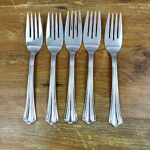 May include: Five stainless steel forks with a decorative, fluted handle design. The forks are arranged in a row on a wooden surface. Each fork has four tines and a detailed, scalloped base.