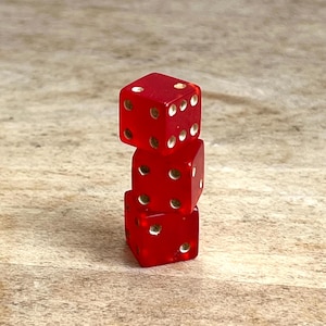 May include: Three red dice stacked on top of each other. Each die has white dots indicating the numbers. The dice are on a light brown wooden surface.