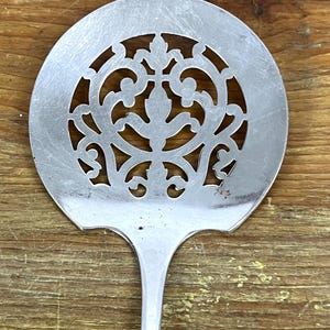 Puede incluir: Una cuchara ranurada de plata con un diseño decorativo y ornamentado en el centro de la cabeza de la cuchara. La cuchara tiene un mango largo y delgado.
