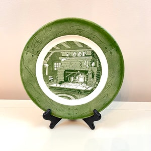 May include: A green and white decorative plate featuring a cabin scene. The plate has a green border with a textured design. The center of the plate depicts a detailed illustration of a cabin interior in shades of green.