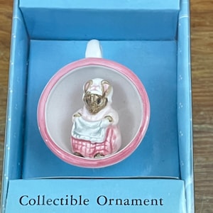 Puede incluir: Adorno coleccionable de Beatrix Potter, presentado en una caja azul claro. El adorno presenta un personaje con un atuendo rosa dentro de un círculo con borde rosa. La caja lleva la etiqueta "Collectible Ornament".
