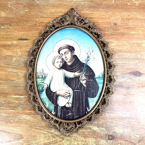 May include: A framed oval print of Saint Anthony of Padua holding a baby Jesus. The print is on a blue background with white lilies and a green landscape in the background. The frame is gold and ornate.