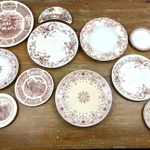May include: A collection of antique brown and white ceramic plates and serving dishes with various floral, landscape, and nautical designs. The plates are arranged on a wooden surface, showcasing their intricate patterns and vintage aesthetic.