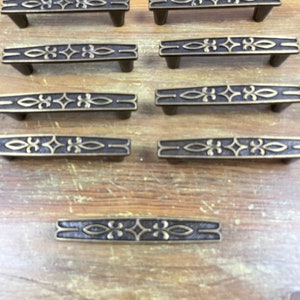 May include: A set of 10 antique brass cabinet pulls with a decorative design. The pulls are all the same size and shape, and they have a slightly aged finish.