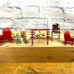 May include: A collection of miniature teddy bear ornaments. The ornaments include a red train, a green bicycle, a red bench, a metal chair, and a red stroller. Each ornament features a teddy bear. The ornaments are displayed on a wooden box.