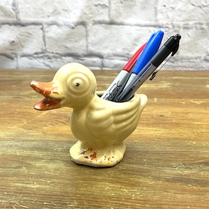 May include: A ceramic duck-shaped pen holder in a pale yellow hue, featuring an orange beak and details. It holds a red, blue, and black Sharpie marker. The pen holder rests on a wooden surface.