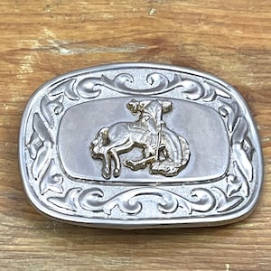 May include: Silver-toned metal belt buckle with a gold-colored rodeo scene. The buckle has an oval shape with a decorative scroll design around the edges. The rodeo scene features a rider on a bucking bronco. A classic Western accessory.