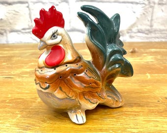 May include: A ceramic rooster figurine with a red comb and wattle, brown and tan body, and green tail feathers. The rooster has small holes on its body and is set on a wooden surface.