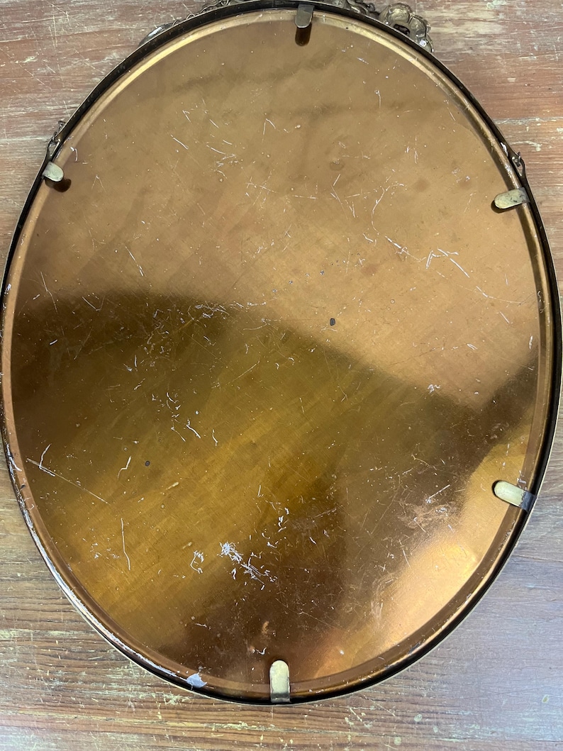 May include: The back of a round, oval-shaped, gold-colored metal picture frame with a smooth surface. The frame has four small, metal hooks on the back.
