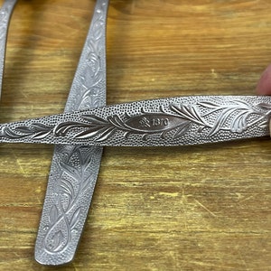 May include: Close-up of several silver spoons with ornate floral patterns. The spoons have a shiny finish and are arranged on a wooden surface. The handle of one spoon displays the text "1870".