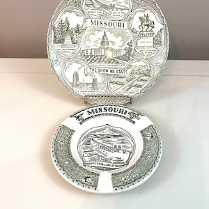 Puede incluir: Dos platos de cerámica vintage con un diseño verde y blanco que presenta imágenes y texto sobre Misuri. El plato más grande tiene un borde de uvas y hojas. El plato más pequeño es un platillo con un diseño de una presa y un lago. Ambos platos tienen la palabra "Missouri" impresa.