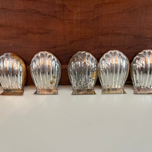 May include: Five silver-colored metal molds shaped like seashells. The molds are arranged in a row on a white surface.