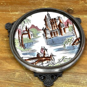 May include: Round metal trivet with a detailed ceramic insert. The insert features a landscape scene with a castle, figures, and trees in shades of brown, blue, green, and pink. The trivet has decorative handles and a base.