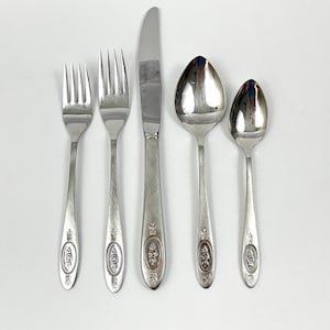 May include: A collection of five silver-coloured cutlery items: two forks, a knife, and two spoons. Each piece has a decorative oval design on the handle. The knife blade is smooth and curved. The spoons are different sizes.