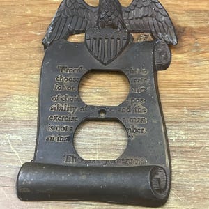 Puede incluir: Una cubierta de enchufe eléctrica de color bronce con un águila posada sobre un pergamino. El pergamino tiene texto de Thomas Jefferson. La cubierta tiene dos aberturas circulares para los enchufes.