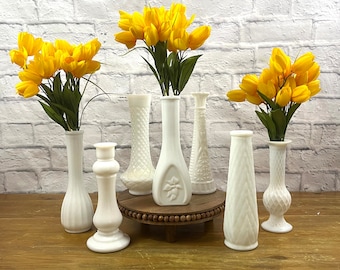 Puede incluir: Una colección de jarrones de vidrio lechoso blanco de varias formas, algunos con tulipanes amarillos brillantes. Los jarrones están dispuestos sobre una superficie de madera, con una pared de ladrillo blanco al fondo.