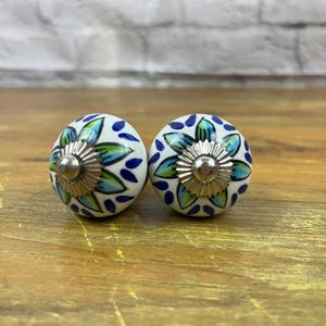 May include: Two white ceramic knobs with blue and green floral designs. The knobs have a silver metal center.