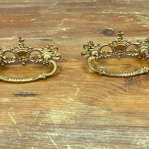 Könnte beinhalten: Zwei verzierte goldfarbene Schubladenknöpfe mit einem dekorativen Schnörkeldesign.