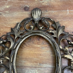 May include: An ornate gold-toned metal picture frame with an oval opening. The frame has a decorative design with intricate scrollwork and floral motifs.