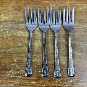 May include: Four silver forks with ornate designs on the handles. The forks are arranged in a row on a wooden surface.