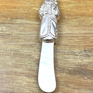 May include: Silver-colored butter knife with a Santa Claus figure on the handle. The Santa figure is detailed with a beard, hat, and coat. The blade is a simple, curved shape.