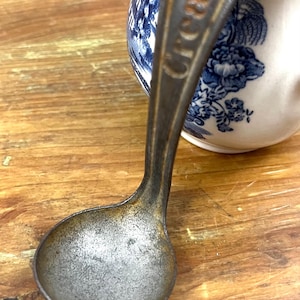 May include: Vintage metal ladle with the word "Cream Top" embossed on the handle. The ladle has a rounded bowl and a long, curved handle. A blue and white floral patterned creamer is in the background.