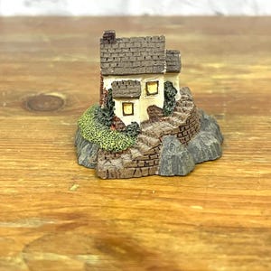 Op de afbeelding: Een klein wit huisje met een bruin dak en twee ramen staat op een rotsachtige basis met groen gebladerte. Het huisje is een miniatuurmodel van keramiek.