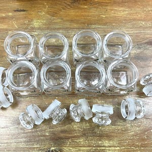 May include: A collection of eight clear glass square jars with matching frosted glass stoppers. The jars are arranged in two rows, with the stoppers scattered around them on a wooden surface. The jars have a simple, classic design.