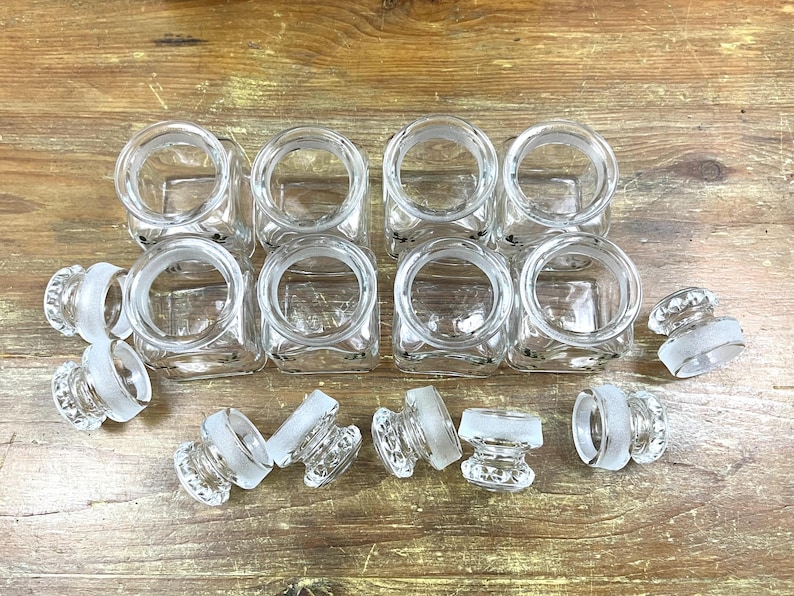 May include: A collection of eight clear glass square jars with matching frosted glass stoppers. The jars are arranged in two rows, with the stoppers scattered around them on a wooden surface. The jars have a simple, classic design.
