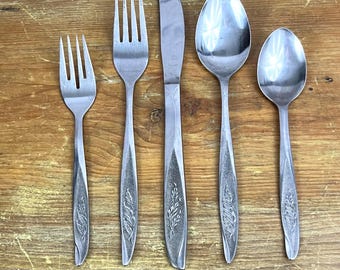 Op de afbeelding: Een set van vijf roestvrijstalen bestekonderdelen: een vork, een mes, een lepel en een kleinere lepel. De handvatten hebben een bladerenpatroon. Het bestek ligt op een houten oppervlak.