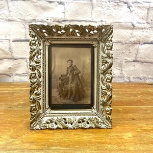 May include: Ornate, vintage picture frame with a sepia-toned portrait. The frame has a gold and white finish with detailed floral and scrollwork. The portrait features a person in period clothing. The frame is displayed on a wooden surface.