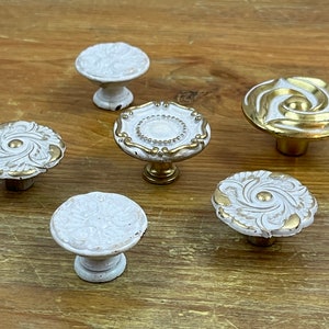 May include: Six decorative cabinet knobs with ornate designs. The knobs are white with gold accents and have a round shape.