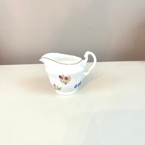 May include: White porcelain creamer with a gold rim and handle. The creamer features a floral design in shades of purple, yellow, and blue. The creamer is a classic piece for serving cream or milk.