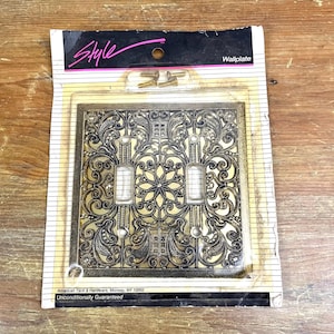 May include: A gold-colored wall plate with a decorative, ornate design. The plate is packaged in a clear plastic bag with a black and white label that reads "Style" and "Wallplate". The label also includes the text "American Tack & Hardware, Monsey, NY 10952" and "Unconditionally Guaranteed".