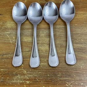 May include: Four silver spoons with beaded edges, arranged in a row on a wooden surface.