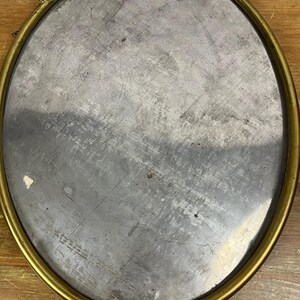 May include: A round, oval-shaped mirror with a gold-colored frame. The frame has a decorative design with floral accents. The mirror is reflecting a wooden surface.