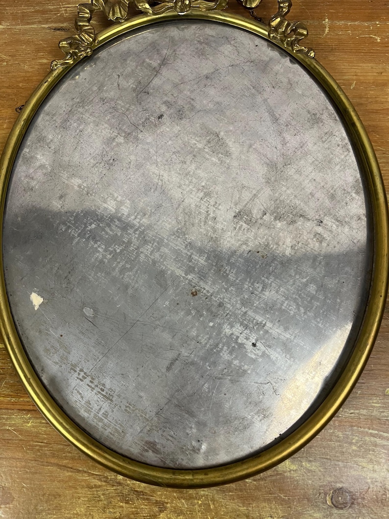 May include: A round, oval-shaped mirror with a gold-colored frame. The frame has a decorative design with floral accents. The mirror is reflecting a wooden surface.