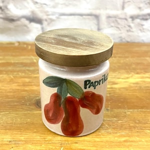 May include: A small, ceramic spice jar with a wooden lid. The jar is beige with a hand-painted design of red peppers and green leaves. The word "Paprika" is written on the jar in black letters. The lid is round and made of light-colored wood.