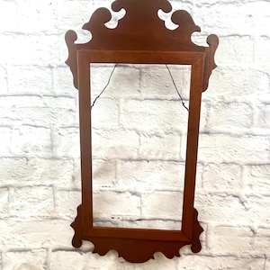 May include: A brown, ornate picture frame with a decorative top and bottom. The frame is rectangular with a dark finish, and is hanging against a white brick wall. The frame has a vintage, antique aesthetic.