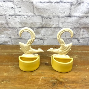 May include: Two ceramic wall pocket planters. Each planter features a cream-coloured swan design with a yellow pocket. The swan's neck curves upwards, and the pocket is a half-circle shape. The planters are on a wooden surface.