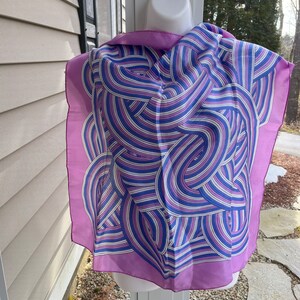 May include: A square silk scarf with a lavender border and a swirling pattern of blue, white, and purple lines. The scarf is displayed on a white mannequin head, showcasing its design and drape.