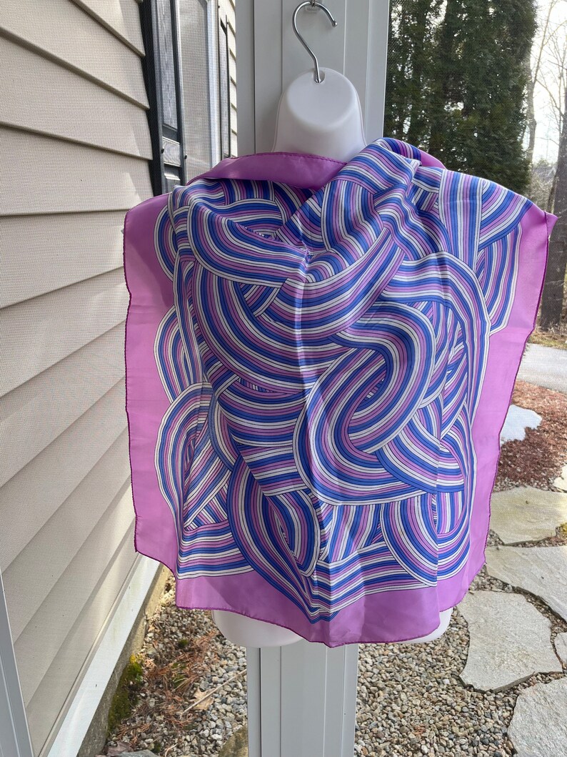 May include: A square silk scarf with a lavender border and a swirling pattern of blue, white, and purple lines. The scarf is displayed on a white mannequin head, showcasing its design and drape.