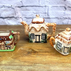 Op de afbeelding: Een set van drie keramische theekopjes in de vorm van een huisje. De theepot is bruin en wit met een groen dak en een bruine handgreep. De roomkan is bruin en wit met een groen dak en een bruine handgreep. De suikerpot is bruin en wit met een bruin dak en een bruine handgreep.